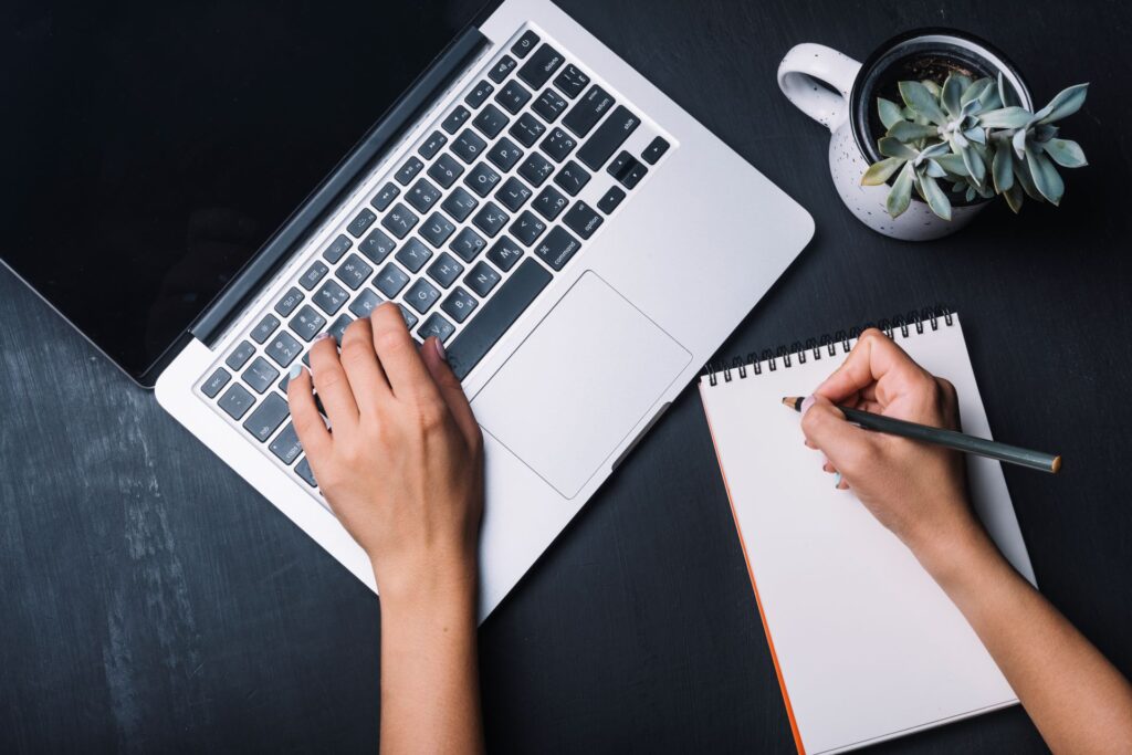 pranayama Overhead flat lay view of a marketer typing on a silver laptop and writing strategy notes on a blank notepad next to a potted plant on a dark desk.