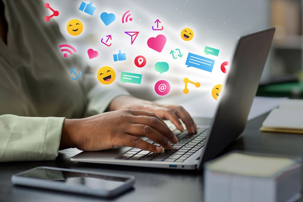 Close-up of a person typing on a laptop with vibrant social media icons like hearts, thumbs-up, smiles, and chat bubbles floating out of the screen.