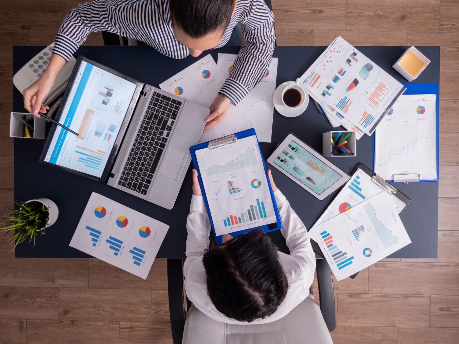 A top-down view of two professionals at a dark grey office desk analyzing multiple printed business reports, charts, and a laptop showing financial data dashboards.