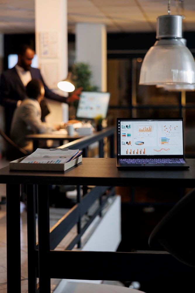 A laptop open on an office desk displaying detailed marketing analytics dashboards, with a blurred team working on strategy in the background under modern lighting.