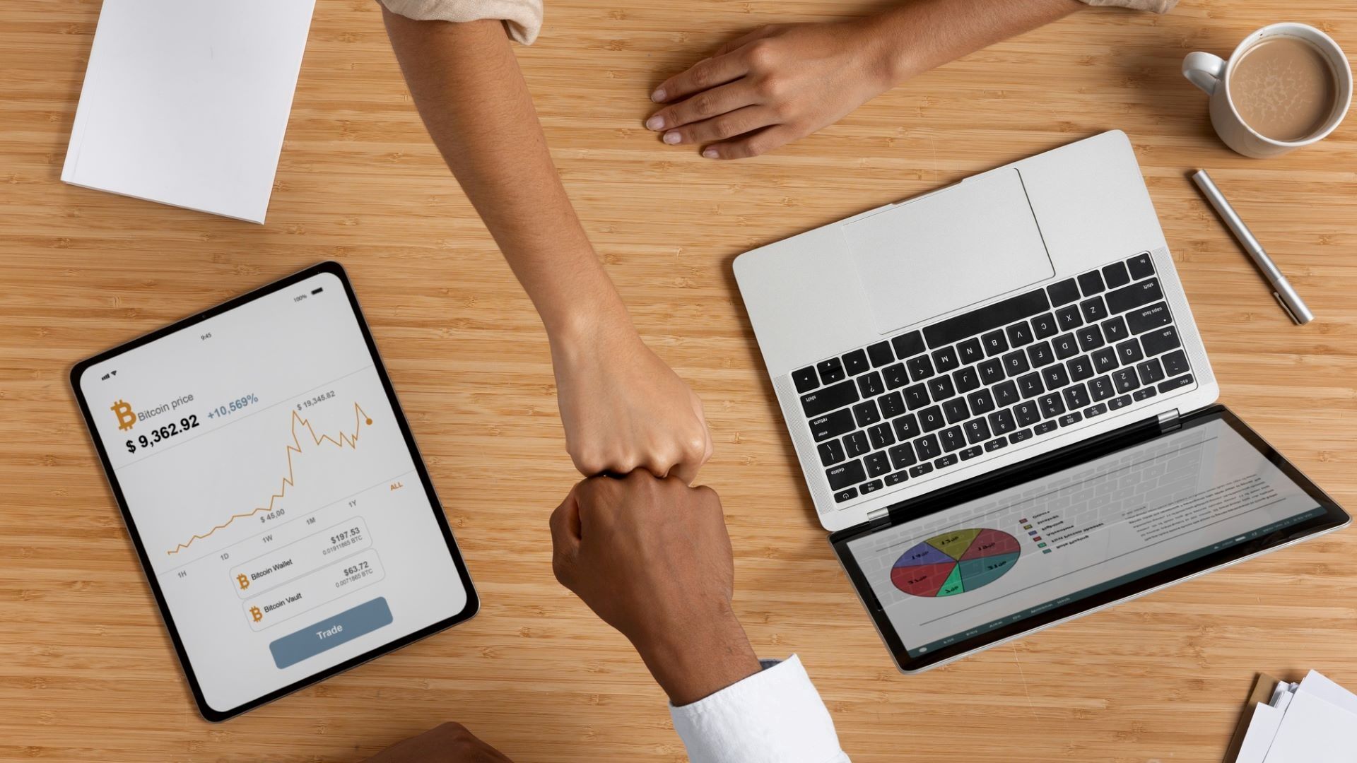 A top-down view of a wooden office desk where a person in white sleeves and a person in a tan shirt are doing a fist bump over an open laptop displaying a colorful pie chart, a tablet showing a Bitcoin price graph, a coffee mug, and a notebook.
